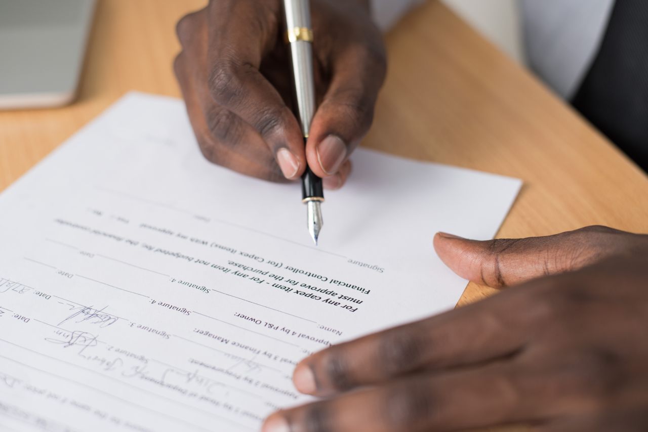 gallery-01 Close-up of a man's hands signing a formal document indoors.